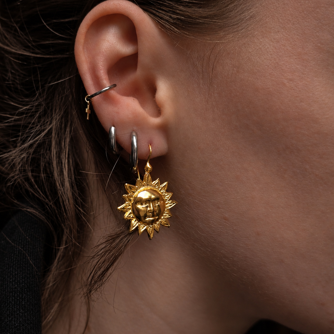 A close-up of a person's ear showcasing a gold sun-face dangle earring, alongside other hoop and cartilage piercings.