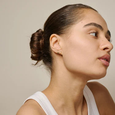 Woman with a bun hairstyle wearing a white top against a beige background ,mini gold plated stud earrings
