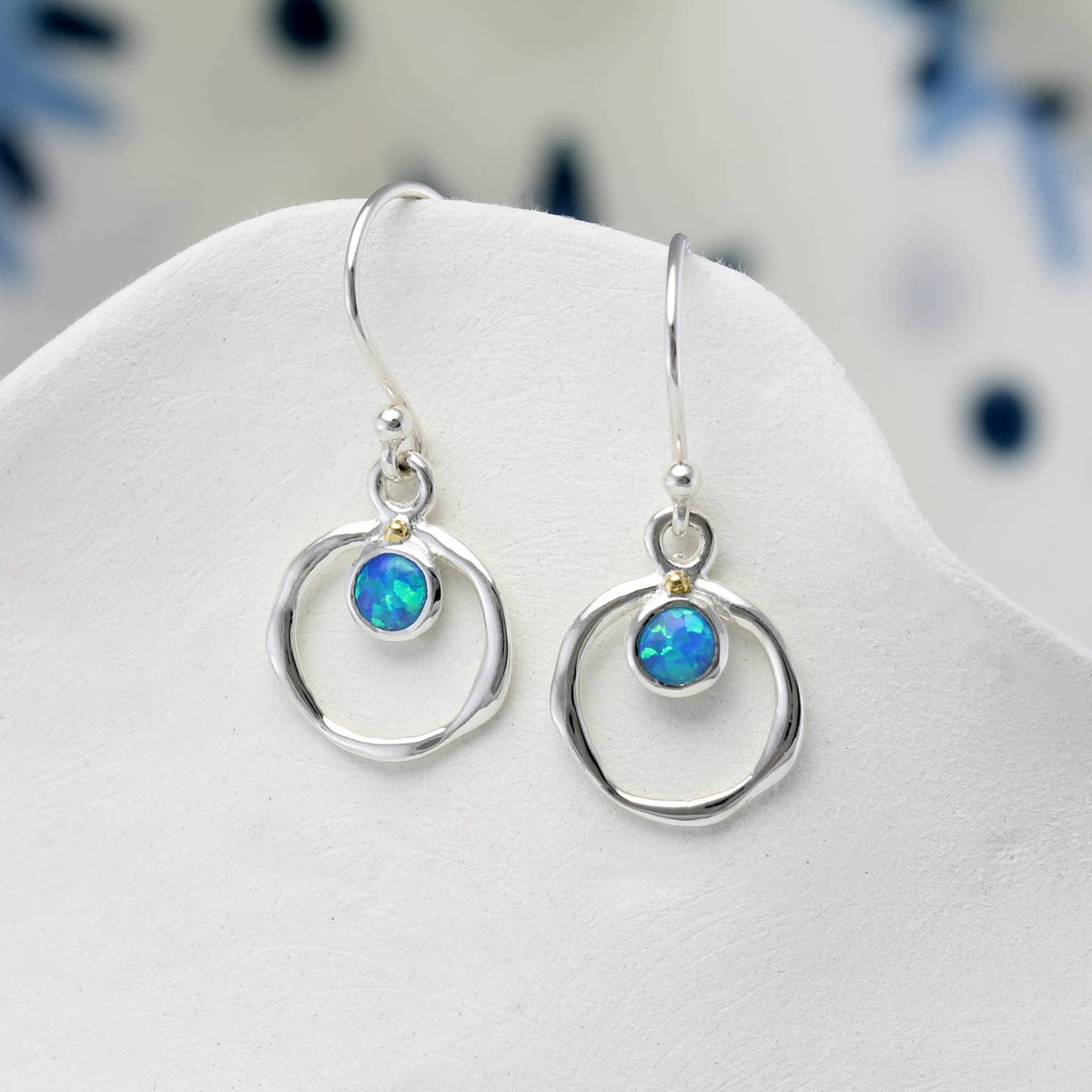A pair of silver dangle earrings with circular wire frames and small blue opal gemstones, displayed on a white jewelry stand.