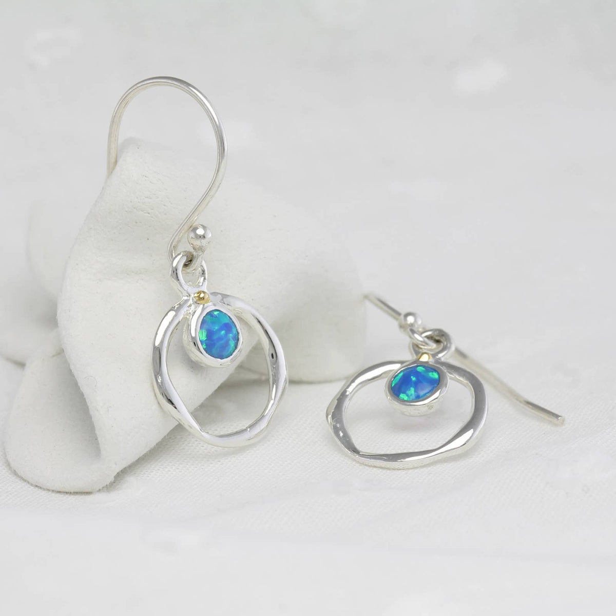 A pair of silver dangle earrings with abstract oval wire frames and small blue opal-like stones, displayed on a soft white fabric surface.