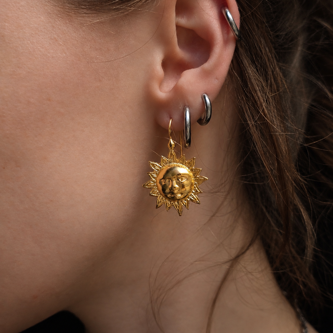 A close-up of a person's ear wearing multiple earrings, including a gold sun-face dangle earring, a silver hoop, and a silver cartilage piercing.
