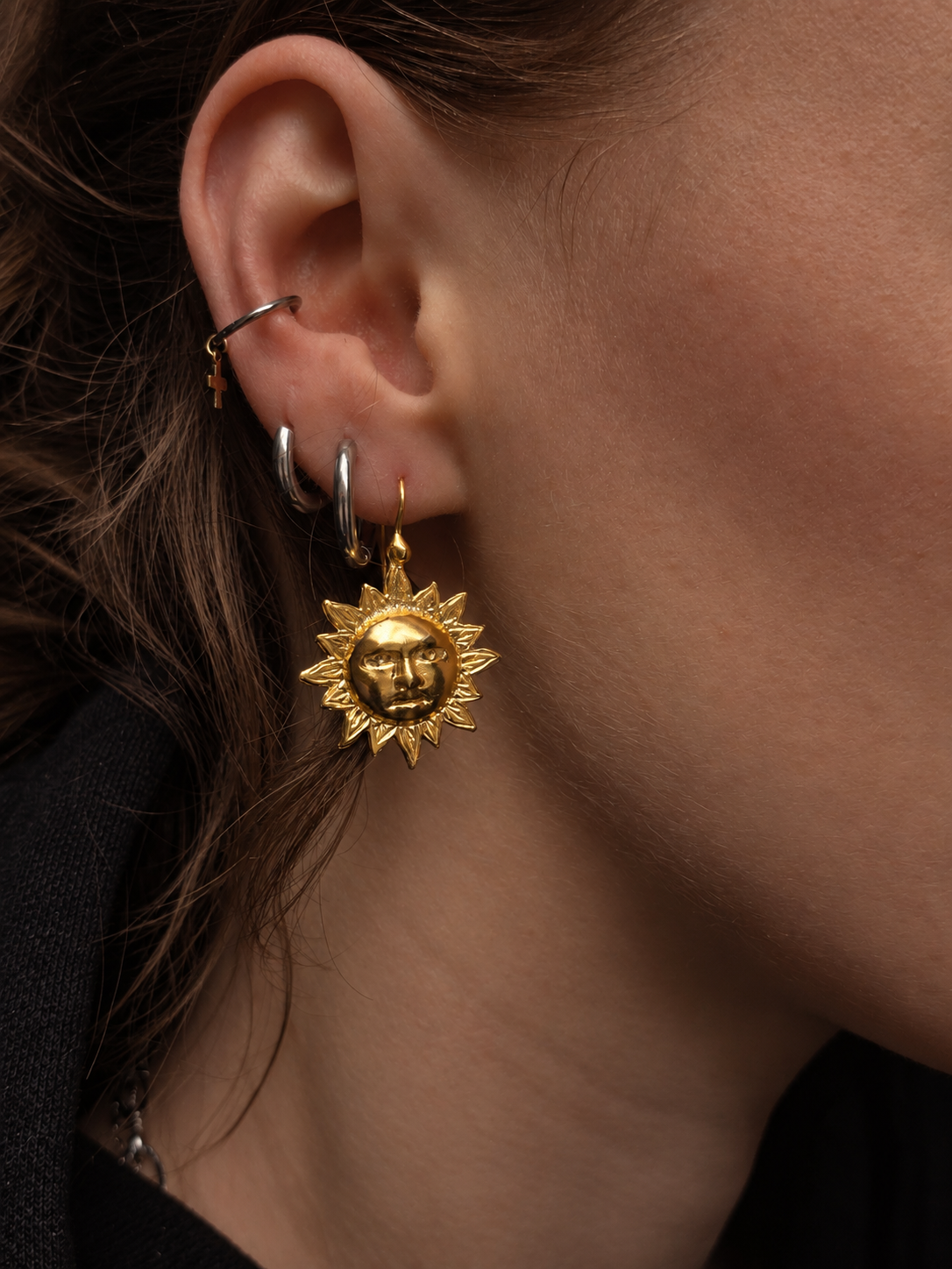 A close-up of a person's ear wearing a gold sun-face dangle earring, along with other hoop and cartilage piercings.